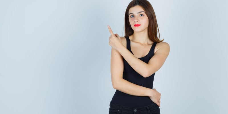 Portrait of beautiful young lady pointing at upper left corner in black tank top and looking confident front view