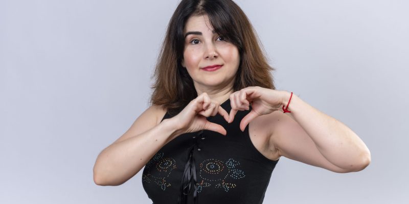 pretty woman in black blouse looking at camera showing her love with fingers standing over white background